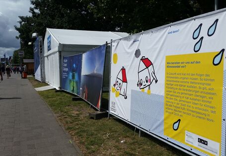 Das Projekt Banner sichtbar zur Straße hin auf der Kieler Woche 2017. Fußgänger laufen auf dem Gehweg. Auf dem Banner sind zwei Zeichnungen von Häusern mit Gesichtern zu sehen. Der Projekttext steht in einer gelben Textbox mit der Überschrift: Wie bereiten wir uns auf den Klimawandel vor.