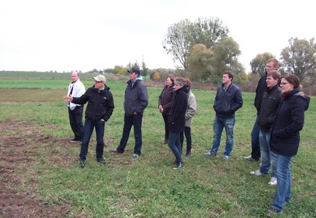 Personen stehen auf einem Rasen schauend in Richtung von Versuchsfeldern und hören dem Fachreferenten zu.  Im Hintergrund befinden sich wenig belaubte Bäume. Der Himmel ist grau.