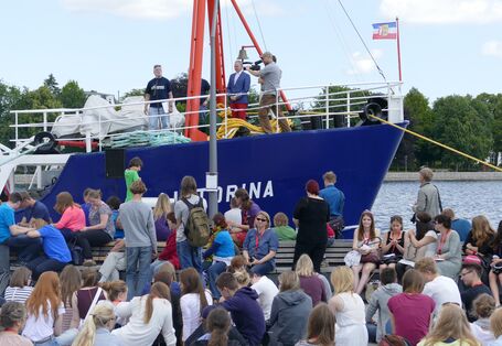 Menschenmenge vor einem Schiff, auf dem Redner und Kameramann stehen.