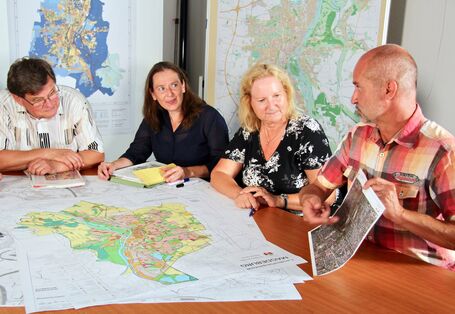 An einem Tisch, auf dem mehrere Karten liegen, sitzen mehrere Personen. Eine Person deutet auf eine kleineren Karte, die sie in der Hand hält.
