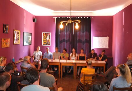 An einer Kopfseite eines Raumes sitzen mehrere Personen in einer Reihe an einem Tisch. Vor ihnen stehen Namensschilder. Ihnen gegenüber sitzen weitere Personen eher ungeordnet im Raum.