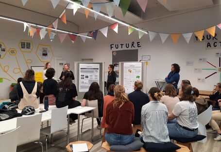 Posterpräsentation im mit Wimpeln ausgestatteten Raum. Der Blick geht über die sitzenden Teilnehmenden hinweg.