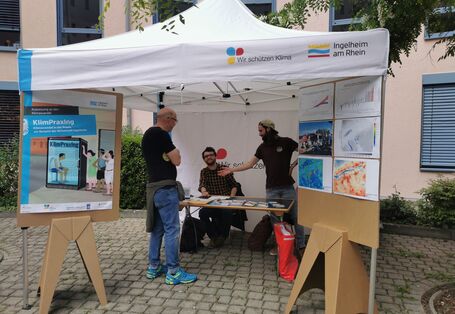 Informationsstand KlimPraxIng mit weißem Dach aus Tuch. Darunter stehen Personen Broschürentisch.