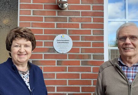 Die zwei Hausbewohner vor ihrem Haus mit einer Plakette für die Auszeichnung als Zukunftshaus an der Wand.