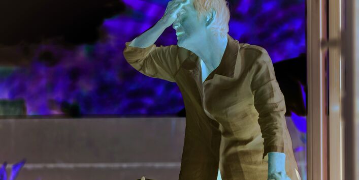 Das Bild zeigt eine ältere Frau mit weißer Bluse. Die Dame hält sich den Handrücken an die Stirn. Im Hintergrund ist sind verschwommen Baumkronen oder Sträucher in hellem Licht zu sehen.