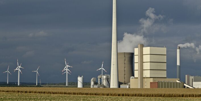 Kraftwerk mit rauchenden Schornsteinen und Windkraftanlagen vor dunkelgrauem Himmel auf einem Feld