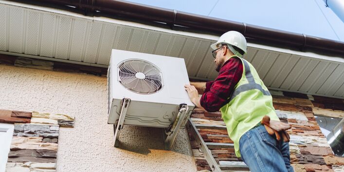 A technician is standing on a ladder and servicing a heat pump.