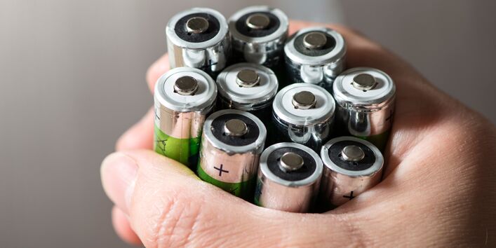 Eine Hand hält ein Bündel gebrauchter AA-Batterien fest umschlossen. Die Pole der Batterien zeigen nach oben, das Gehäuse einiger Batterien ist grün-silberfarben.