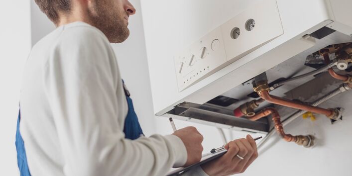 Ein Handwerker in blauer Arbeitskleidung und weißem Pullover steht vor einem geöffneten Boiler und notiert sich etwas auf einem Klemmbrett.