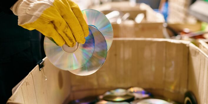 Eine Hand in einem gelben Arbeitshandschuh hält mehrere glänzende CDs über einen großen Karton, der bereits mit vielen weiteren CDs und DVDs gefüllt ist.