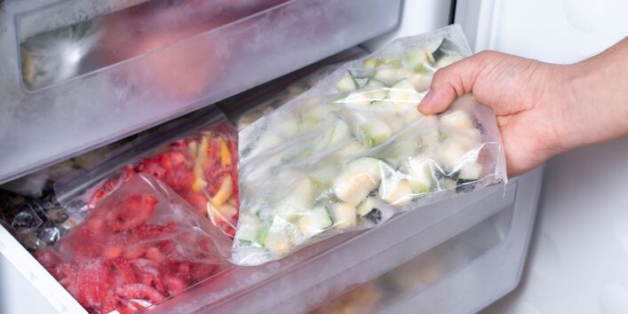 Eine Hand holt eine Plastiktüte mit gefrorenen Zucchini-Stücken aus dem Gefrierfach eines Kühlschranks. Im Fach darunter befinden sich Tüten mit anderen gefrorenen Lebensmitteln, wie zum Beispiel Paprika.