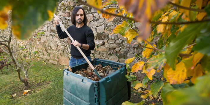 Ein Mann mit dunklen Haaren und Bart in einem schwarzen Pullover und blauen Jeans steht im Garten vor einer grünen Komposttonne und mischt den Inhalt mit einem Rechen um. Im Hintergrund ist eine Natursteinmauer zu sehen und im Vordergrund hängt ein Ast mit gelben und grünen Blättern.