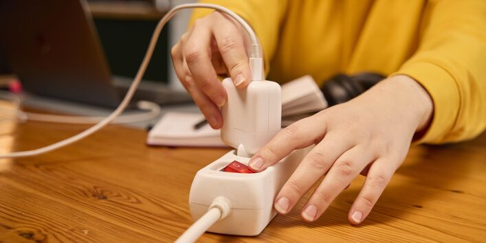 Eine Person steckt ein weißes Ladekabel in eine weiße Steckerleiste, während der rote Ausschalter daneben leuchtet. Im Hintergrund sind verschwommen ein Laptop und Kopfhörer zu sehen.