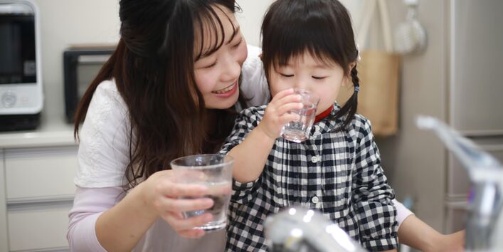 Eine lächelnde Frau beobachtet in einer Küche ein kleines Mädchen, das konzentriert aus einem Glas frisches Leitungswasser trinkt. Im Vordergrund ist ein moderner Wasserhahn zu sehen.