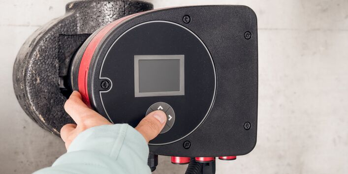 Eine Hand drückt auf einen Einstellknopf einer Heizungspumpe mit einem digitalen Display.