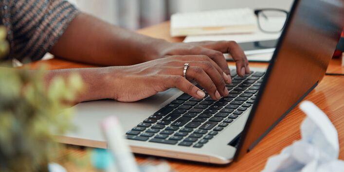 Eine Person mit einem silbernen Ring an der Hand tippt auf einer Laptoptastatur. Im Hintergrund sind verschwommen eine Brille, ein Stift und zerknülltes Papier zu sehen. 