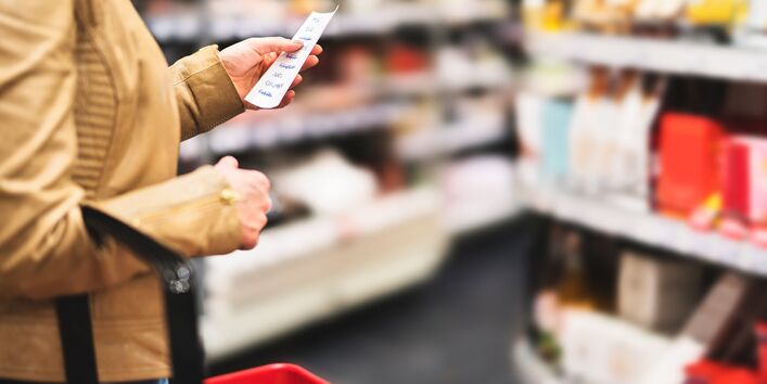 Eine Person in einer beigen Jacke und mit einem roten Einkaufskorb in der Hand hält in einem Supermarkt eine Einkaufsliste in die Höhe und blickt darauf.