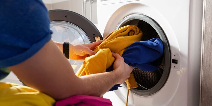 Eine Person steckt bunte Wäsche in eine Waschmaschine. Die offene Tür der weißen Waschmaschine ist zu sehen. Im Vordergrund liegt ein Haufen bunter Kleidung.
