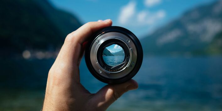 Im Vordergrund ist eine Hand zu sehen, die ein Objektiv hält. Im Hintergrund ist eine Bergsee-Landschaft zu sehen, das Objektiv fokussiert in den Horizont hinein.