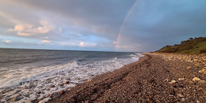 Wellen der Ostsee schlagen an einen Steinstrand, der in grüne Hügel übergeht. Der Himmel ist über den Hügeln graublau aber über dem Meer hellblau mit weißen Wolken. Ein Regenbogen beginnt über dem Meer.  