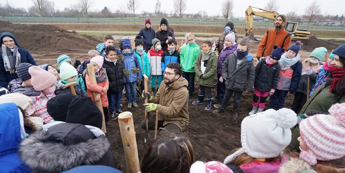 Kinder in Winterkleidung lernen von einem Erwachsenen im Freien das Pflanzen eines Baumes. Im Hintergrund ist ein Bagger zu sehen.
