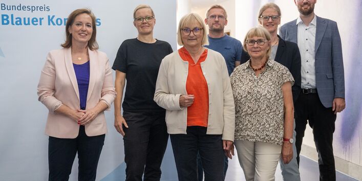 Auf dem Bild sind die Jurymitglieder vor dem Blauen Kompass Logo zu sehen. Dr. Cornelia Lawrenz (Stiftung THW e.V.), Nadine Steinbach (Verband kommunaler Unternehmen), Dr. Bettina Hoffmann (BMUV), Toralf Staud (klimafakten.de), Prof. Dr. Andrea Heilmann (Hochschule Harz), Prof. Dr. Lilian Busse (UBA), Sebastian Lummel (Deutscher Städte und Gemeindebund) und nicht auf diesem Bild Annika Roth (Klimaschutz Unternehmen e.V.) als weiteres Jurymitglied.