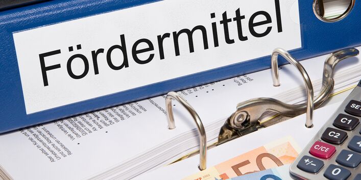 Foto: Aktenordner mit Beschriftung „Fördermittel“, davor auf einem Tisch liegen Geld, Taschenrechner und Kugelschreiber.