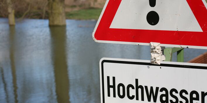 Foto: Ein überschwemmtes Gebiet während eines Hochwasserereignisses. Im Vordergrund steht ein „Achtung Hochwasser“ Schild