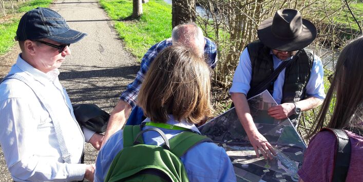 Foto: Mehrere Personen blicken vor einem technisch ausgebauten Fluss auf Planungsunterlagen.
