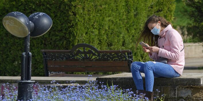 Eine Frau sitzt in einem Park auf einer Bank, trägt einen Mund-Nasen-Schutz und schaut auf ihr Smartphone