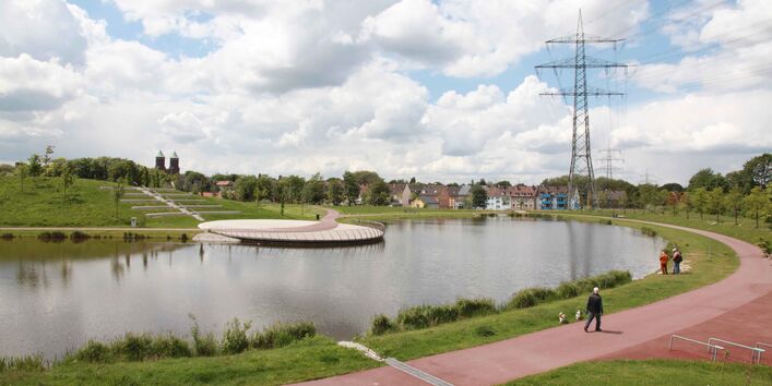 Das Bild zeigt einen angelegten kleinen See mit Grünanlagen und Häusern im Hintergrund