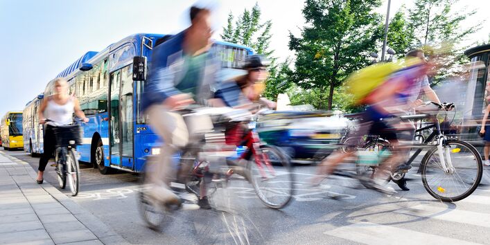 Ein hektisches Verkehrsgeschehen ist abgebildet. Busse und schnell fahrenede Radfahrer sind zu sehen. 