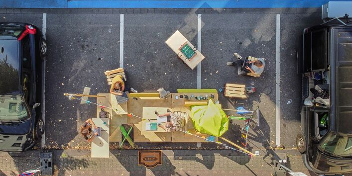 Auf einem Parkplatz steht ein großes Gefährt quer und Menschen sind aus der Vogelperspektive zu sehen, die im Freien an ihren Laptops arbeiten. 