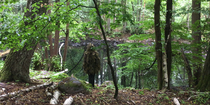 Fotografie eines Menschen, der mit Naturmaterialien verkleidet ist und im Wald vor einem Teich steht