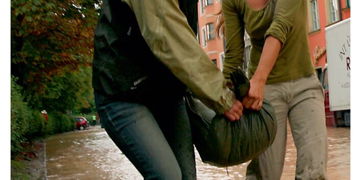 Cover von "Anpassung an den Klimawandel: Hochwasserschutz", Titelfoto: Zwei Frauen tragen gemeinsam einen Sandsack durch eine überflutete Straße