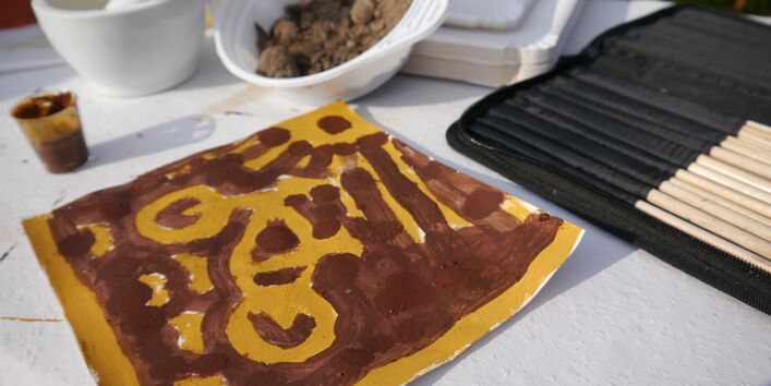 Dargestellt ist ein Fahrrad im Sonnenschein, gemalt mit Bodenfarben von Teilnehmenden der Fortbildung, im Hintergrund der verwendete Boden in einer Schale