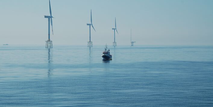 Das Foto zeigt drei Windenergieanlagen im Meer mit Jacket-Fundament (als Fachwerk konstruierte Gründungsstruktur). Ganz im Hintergrund ist die Forschungsplattform FINO1 sichtbar. Im Vordergrund ist ein im Verhältnis zu den Windenergieanlagen klein wirkendes Schiff sichtbar.