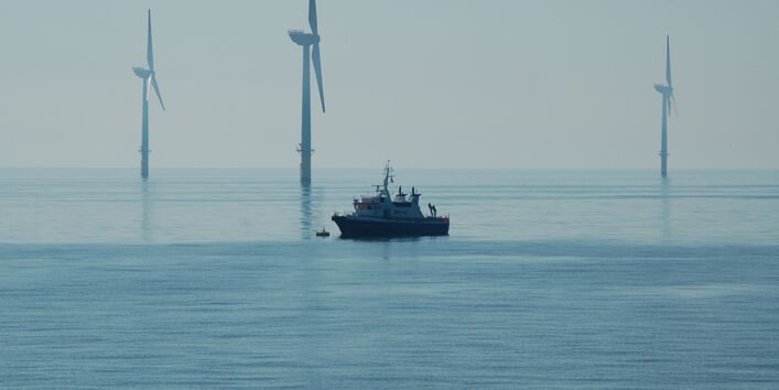 Das Foto zeigt ein Schiff vor drei Windenergieanlagen mit einem Monopile-Fundament. Davor sieht man ein im Verhältnis sehr klein wirkendes Schiff.