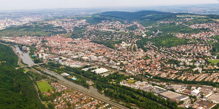 Luftbild einer Gemeinde in hügeliger Landschaft mit Fluss, Autobahn, Gewerbegebiet und Bahnstrecke im Vordergrund