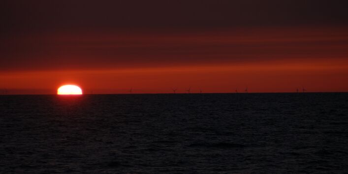Das Foto zeigt einen Sonnenuntergang über dem Meer. Am Horizont sind bei dem Windenergieanlagen zu sehen.