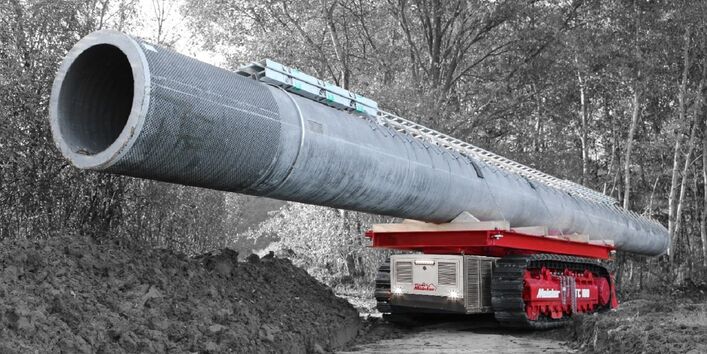 ein kleines selbstfahrendes Raupenfahrzeug transportiert auf seiner Ladefläche einen großen Betonmast durch den Wald