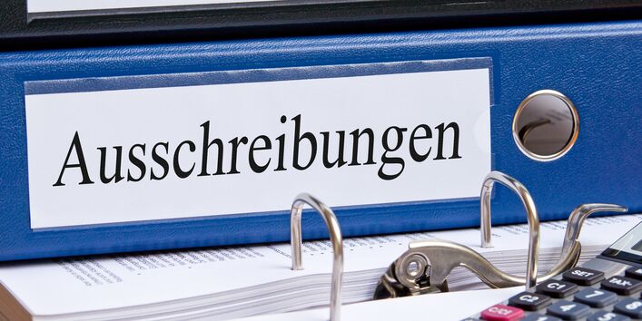 auf einem Tisch liegen ein Ordner mit der Aufschrift "Ausschreibungen", ein Taschenrechner und ein Kugelschreiber