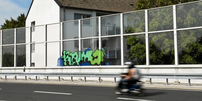 ein Wohnhaus liegt direkt an einer Stadtautobahn hinter einer Lärmschutzwand, gerade fährt ein Motorrad vorbei