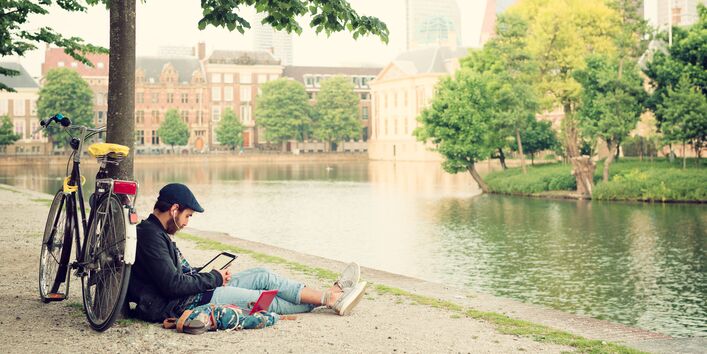 Ein junger Mann sitzt in der Stadt an einem Fluss unter einem Baum und schaut auf sein Tablet mit Kopfhörern im Ohr. Am Baum lehnt sein Fahrrad.