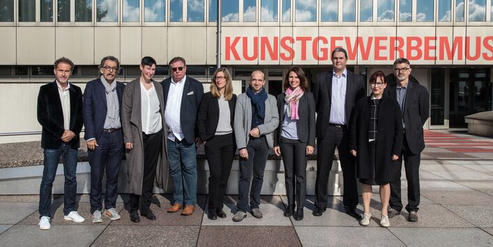 10 Männer und Frauen haben sich vor einem Gebäude mit der Aufschrift "Kunstgewerbemuseum" für ein Gruppenfoto aufgestellt