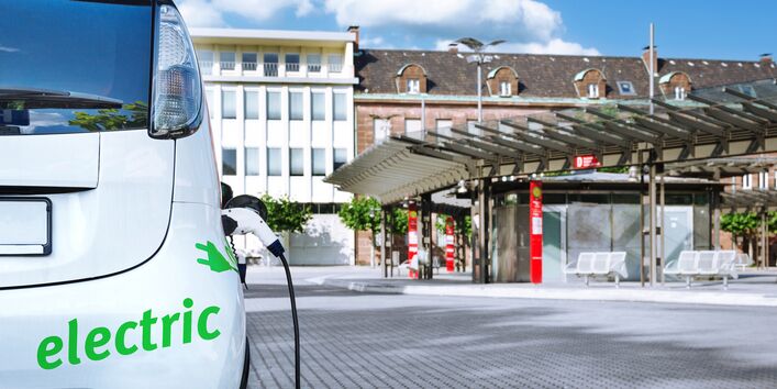 Ein Elektroauto wird aufgeladen, im Hintergrund eine moderne Bushaltestelle in der Stadt