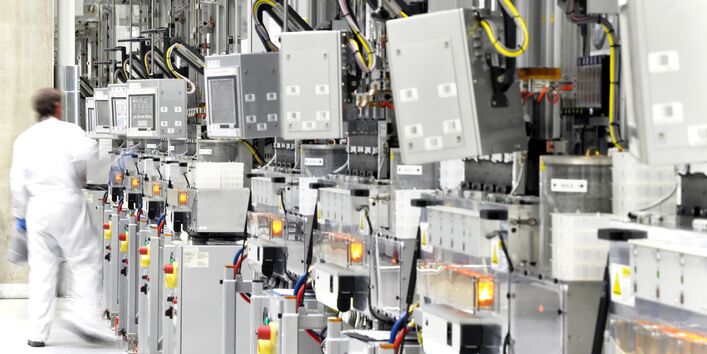 ein Mitarbeiter in einem weißen Schutzanzug in einem Betriebsraum mit vielen elektrischen Geräten, Kabeln und Kästen, die an der Wand stehen oder aufgehängt sind