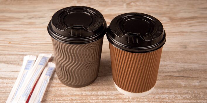 zwei „Coffee to go“-Becher auf einem Tisch, daneben zwei Papiertütchen mit Zucker