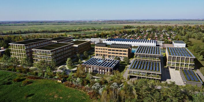 die fotorealistische Computersimulation zeigt ein inmitten von Feldern und Bäumen gelegenes Gelände mit mehreren modernen Gebäuden mit Holzfassaden, Begrünung und Solaranlagen auf den Dächern