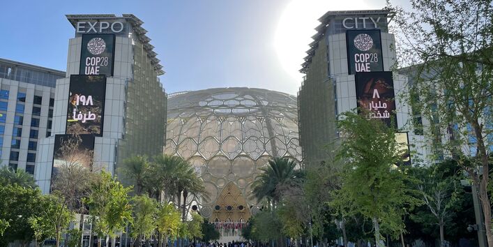 Menschen streben dem Eingang eines modernen Gebäudes mit der Aufschrift Expo City COP 28 UAE zu, auf der Straße sind Bäume in Kübeln als Allee aufgestellt